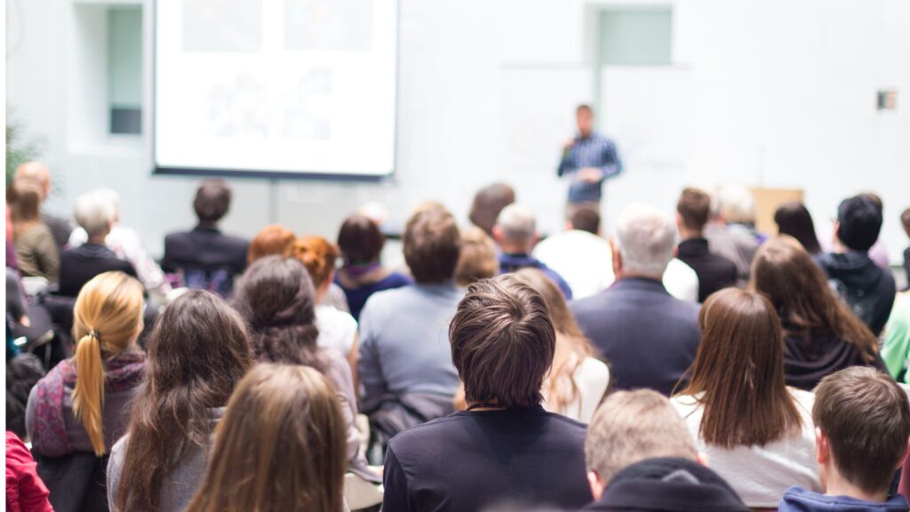 Audience seated in rows watching a speaker presenting in a bright conference room with a projection screen.
