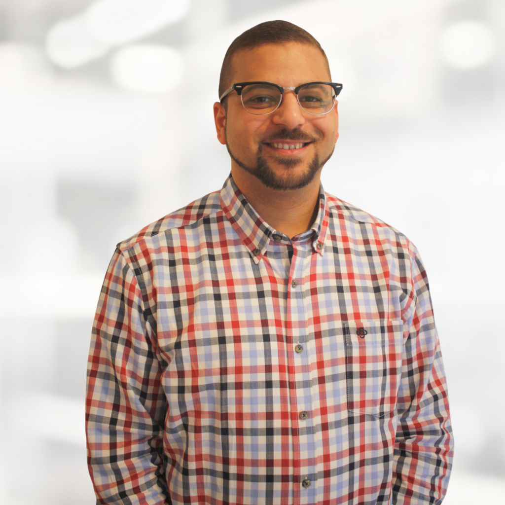 Smiling man wearing a red, white, and blue plaid shirt and glasses against a blurred light background.