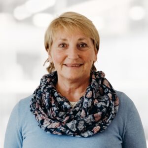 Smiling woman with short blonde hair, wearing a light blue top and a butterfly-patterned scarf, in a bright indoor setting.