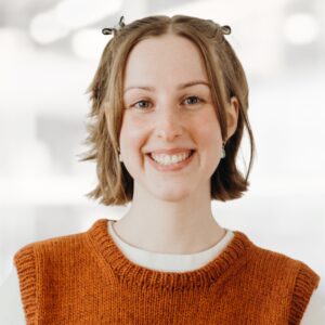 Smiling woman with short brown hair wearing an orange knitted sweater, neutral blurred background.