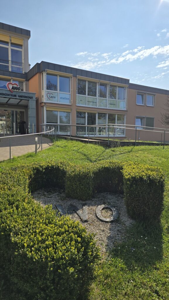 Exterior view of a modern educational building with a ramp-accessible entrance, large windows, and a sign reading 'Lernstation' under a blue sky.