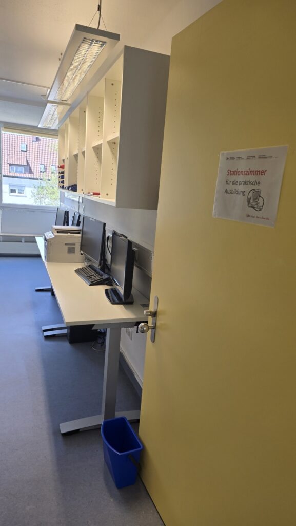 Workstation in a classroom: desk with two computer monitors and a printer, white shelves above, yellow wall to the right, blue waste bin nearby.
