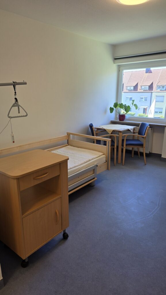 Patient room with wooden single bed, small nightstand, and a dining table by the window; a potted plant sits on the sill.