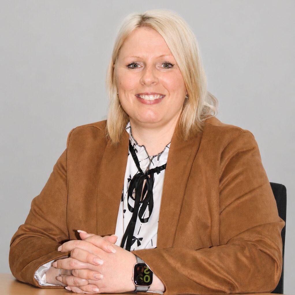Smiling blonde woman in a brown blazer sits at a desk with hands folded, keys in the foreground.