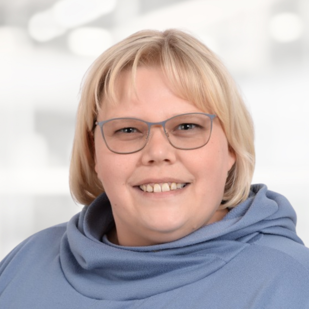 Smiling woman with short blonde hair and glasses wearing a blue hoodie, posed against a soft, blurred indoor background.