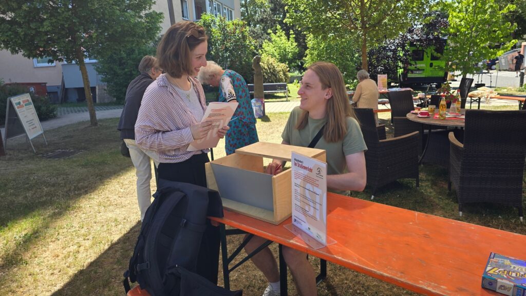 Two women talk at an outdoor information booth, one holding papers and smiling at the other seated behind a wooden box on a bright day.