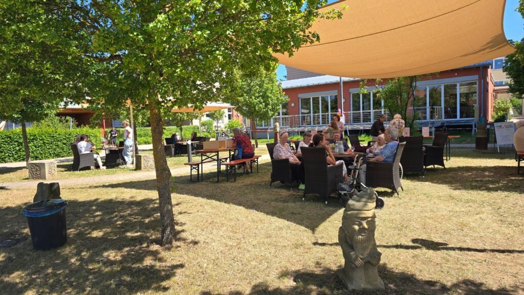 Outdoor café under a beige shade sail with groups of seniors seated at wicker tables, chatting near a red-brick building and leafy trees.