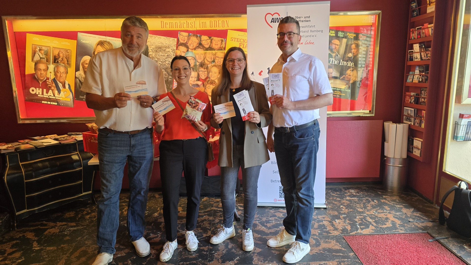 Marie machts_Kinoabend_1 - AWO Kreisverband Bamberg - AWO Kreisverband Bamberg Group of four adults posing with flyers in a movie-themed lobby, red walls, posters, and a banner with a heart logo in the background.