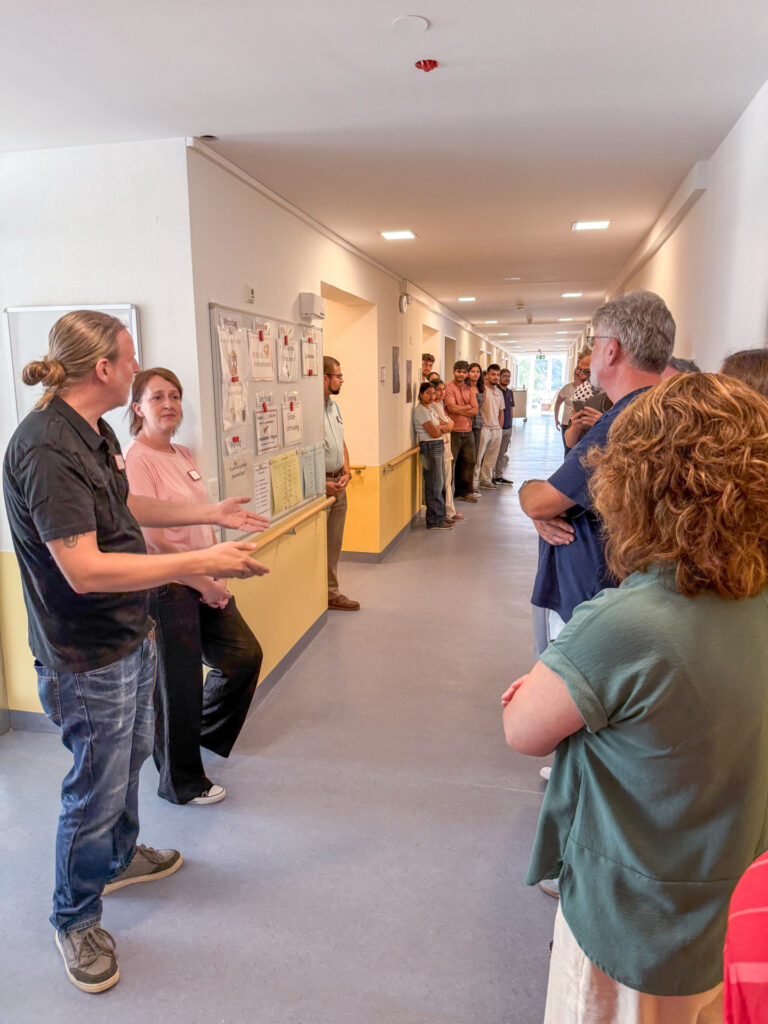 People stand in a long hallway, watching two presenters by a wall with a bulletin board full of notices talk to the crowd.