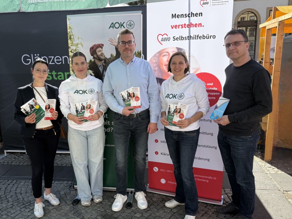 Group of five adults posing for a photo at a health information stand, two wearing white AOK hoodies and holding brochures.
