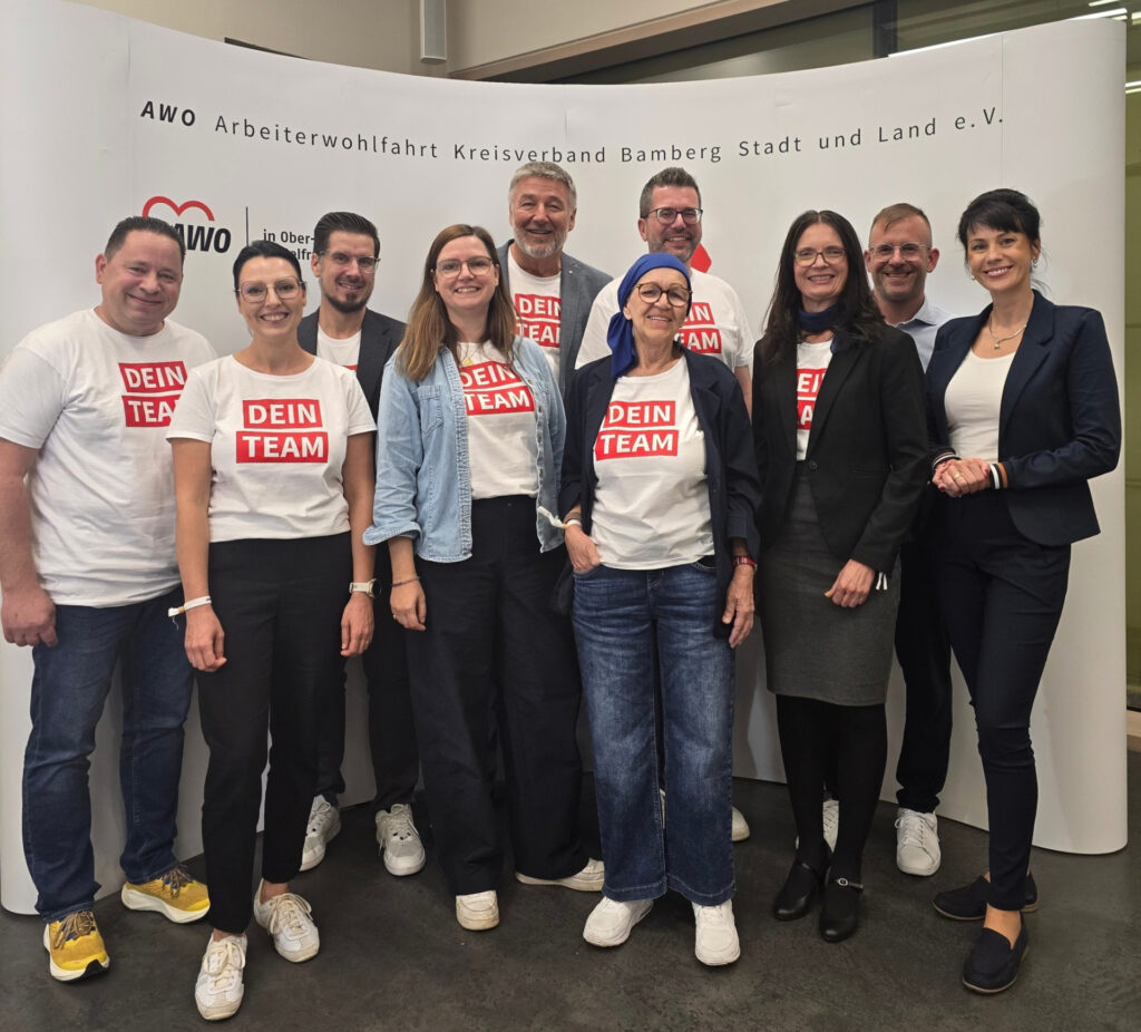 Group of ten adults posing together indoors, several wearing white t-shirts with a red 'DEIN TEAM' logo, smiling at the camera.”,”Team photo of colleagues in casual and business attire, posing in front of a banner.”,”Smiling group of coworkers at an event, standing shoulder-to-shoulder in front of a curved backdrop.”,”Ten people posing for a team photo in an office setting, some wearing matching 'DEIN TEAM' shirts.”,”Diverse group of coworkers posing for a photo at an event, with a backdrop featuring organizational branding.