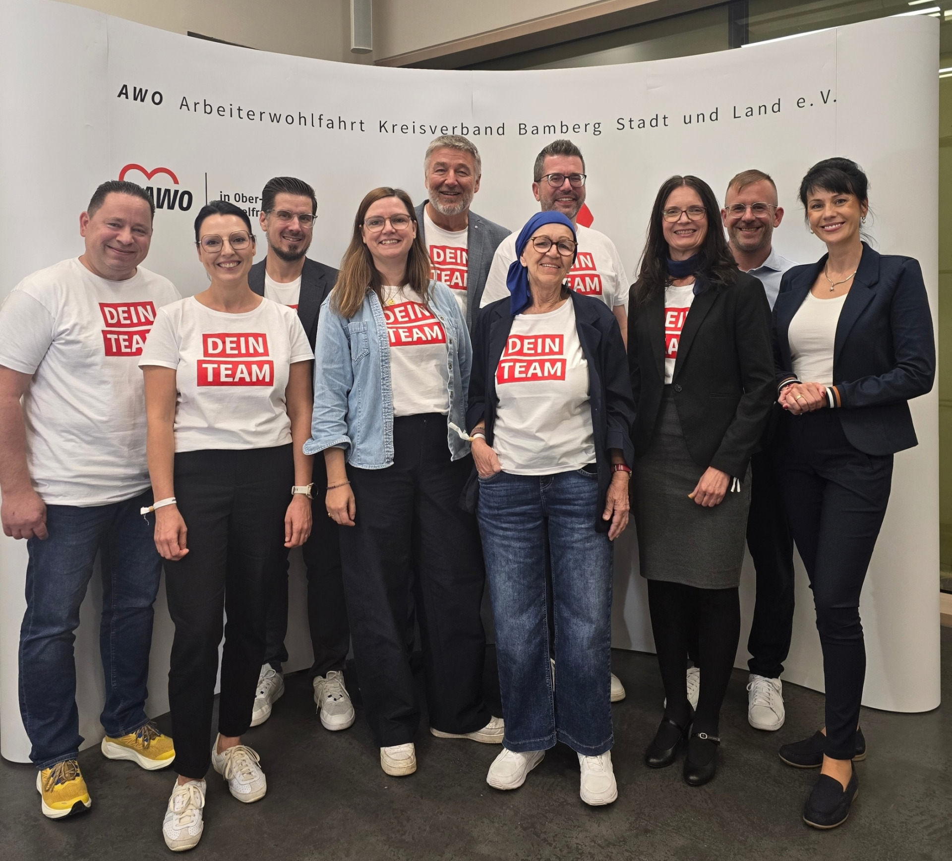 Group of ten adults posing together indoors, several wearing white t-shirts with a red 'DEIN TEAM' logo, smiling at the camera.”,”Team photo of colleagues in casual and business attire, posing in front of a banner.”,”Smiling group of coworkers at an event, standing shoulder-to-shoulder in front of a curved backdrop.”,”Ten people posing for a team photo in an office setting, some wearing matching 'DEIN TEAM' shirts.”,”Diverse group of coworkers posing for a photo at an event, with a backdrop featuring organizational branding.