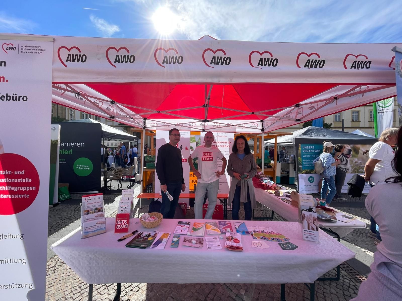 Outdoor information booth at a sunny fair, volunteers stand behind a white-and-red canopy with AWO logos and a table of brochures in front.