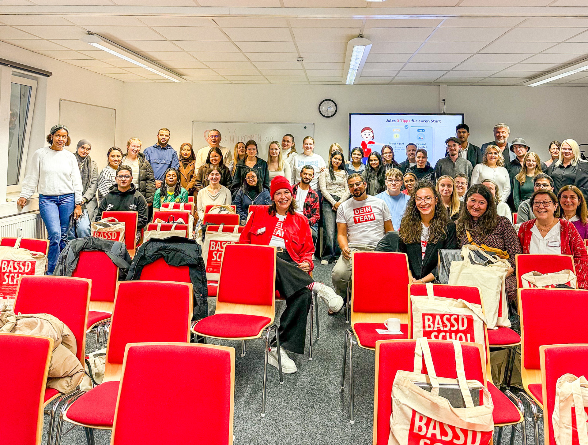 Group of diverse adults posing for a photo in a classroom with red chairs and a presentation screen in the background.
