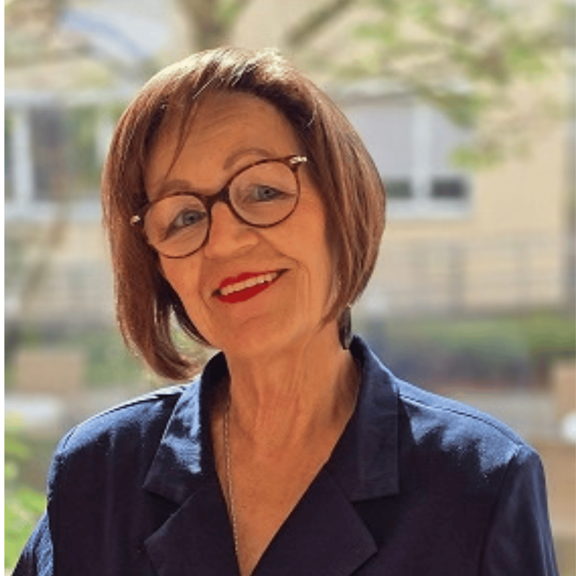 Smiling woman with short brown hair and round glasses wearing a navy blazer outdoors.